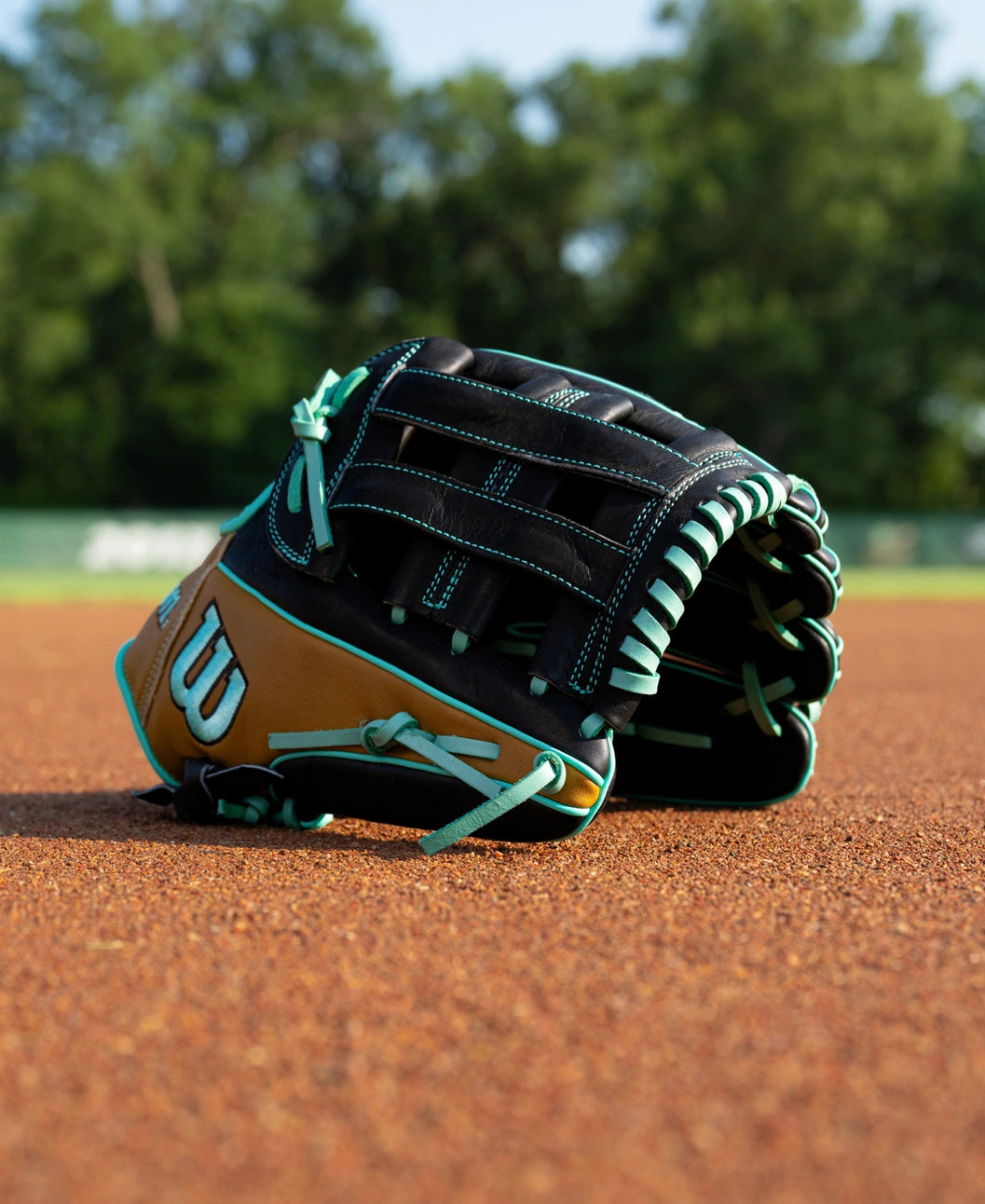 Wilson A1000 DW5 12″ Baseball Glove: WBG02701RTDW5 in tan and black with teal lacing resting on a dirt infield.