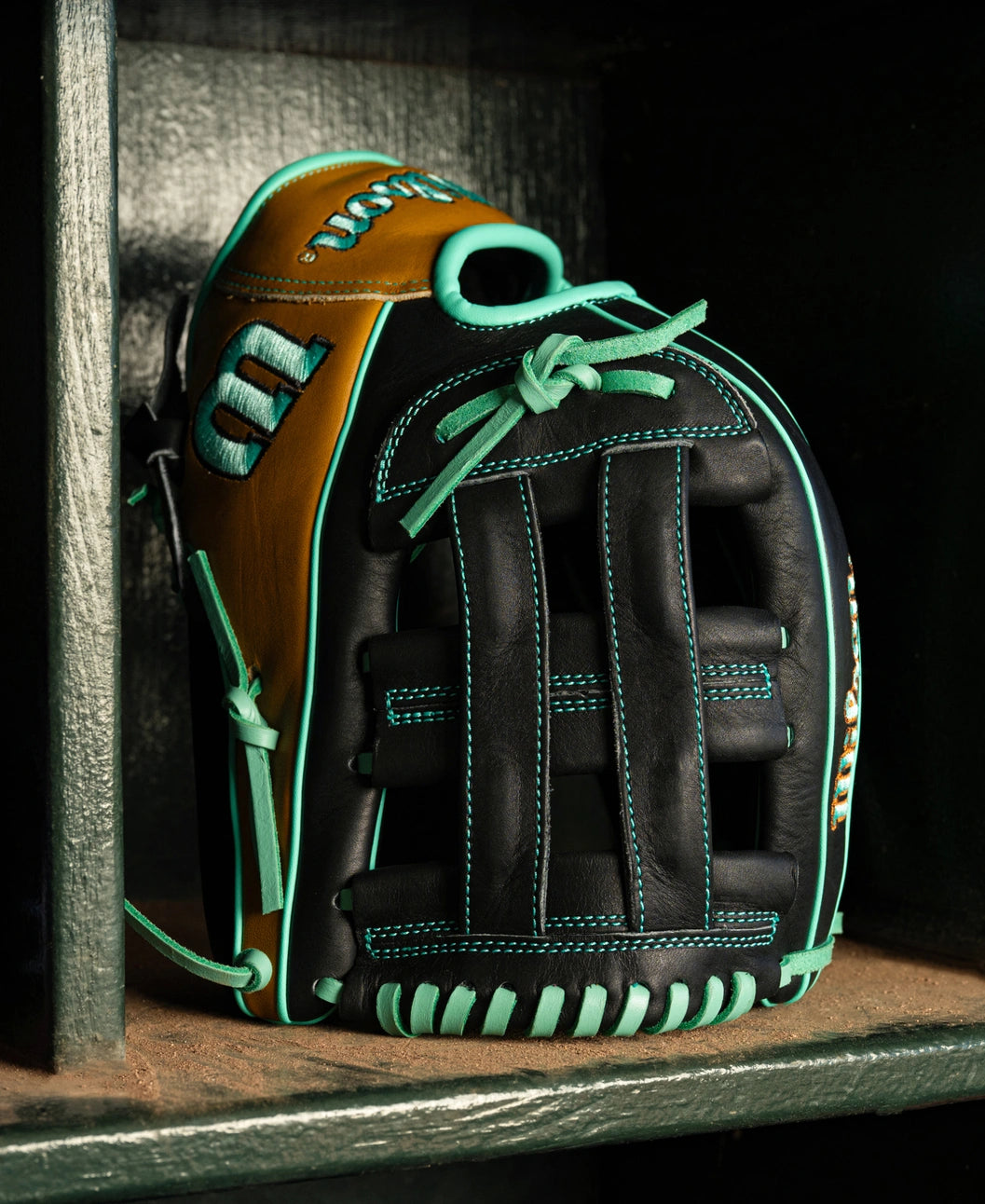 Wilson A1000 DW5 12″ Baseball Glove: WBG02701RTDW5 in black and tan with teal lacing and stitching, sitting on a wooden dugout bench.