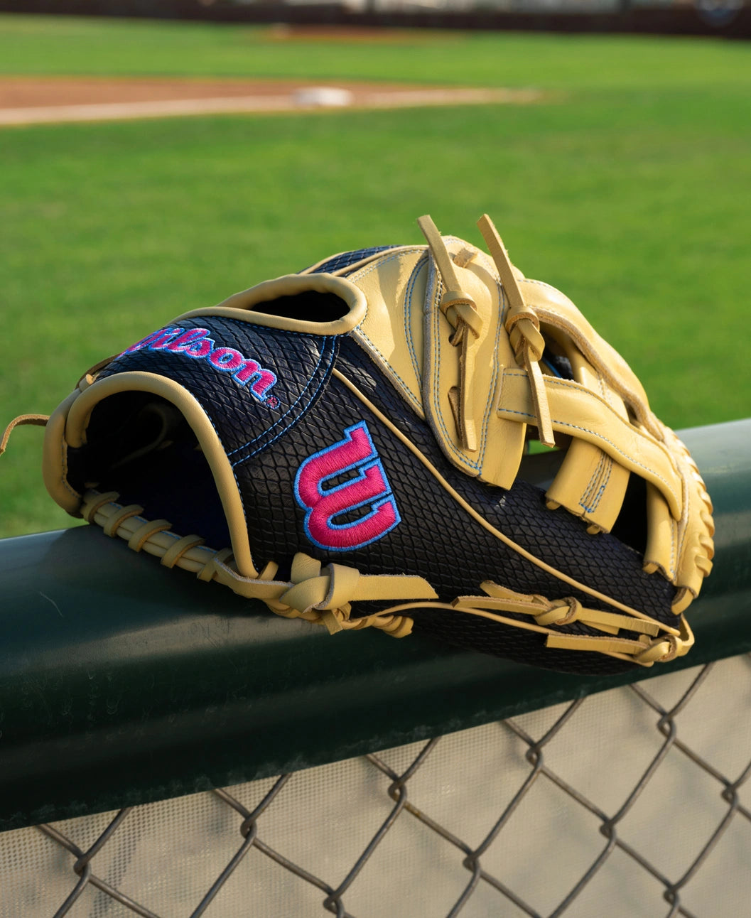 Tan and black Wilson A1000 1750 12.5″ Baseball Glove: WBG02901RT1750 outfield glove with blue and pink logos resting on a chain-link fence in front of a baseball field.