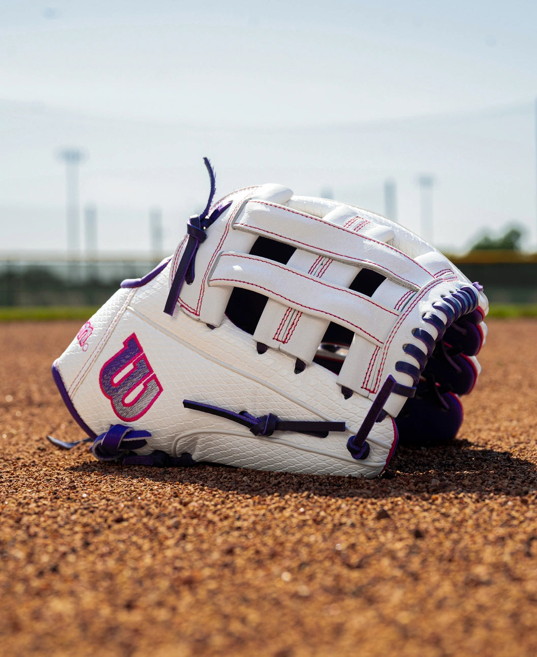 White and purple Wilson A1000 FP1712SS 12″ Fastpitch Glove sitting on a dirt field.