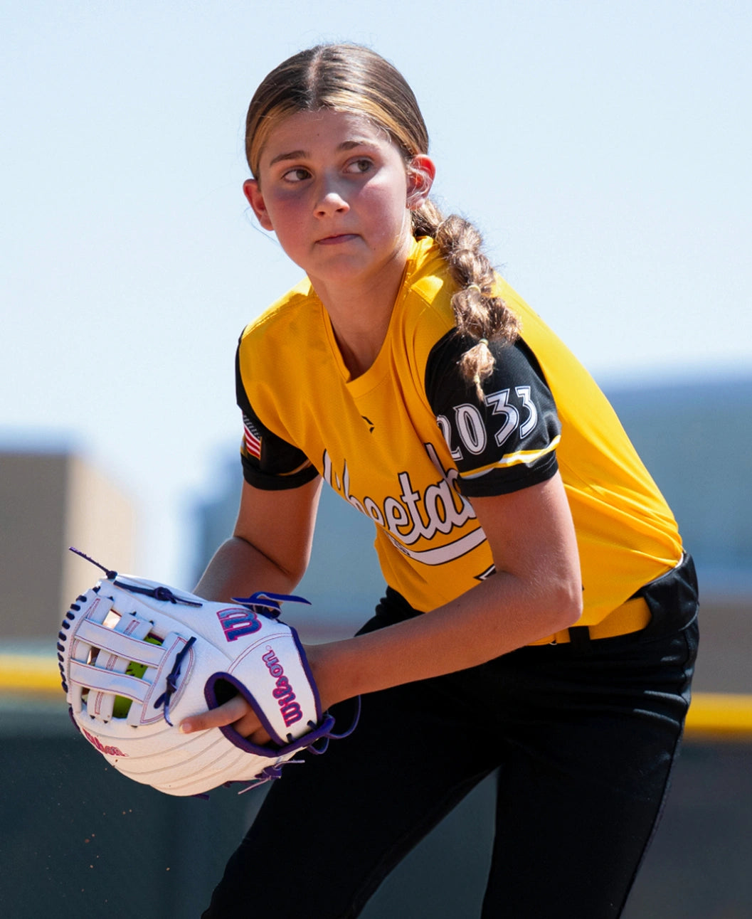 A young softball player in a yellow jersey holds a white Wilson A1000 FP1712SS 12″ Fastpitch Glove with a Dual Post Web while looking toward the field.