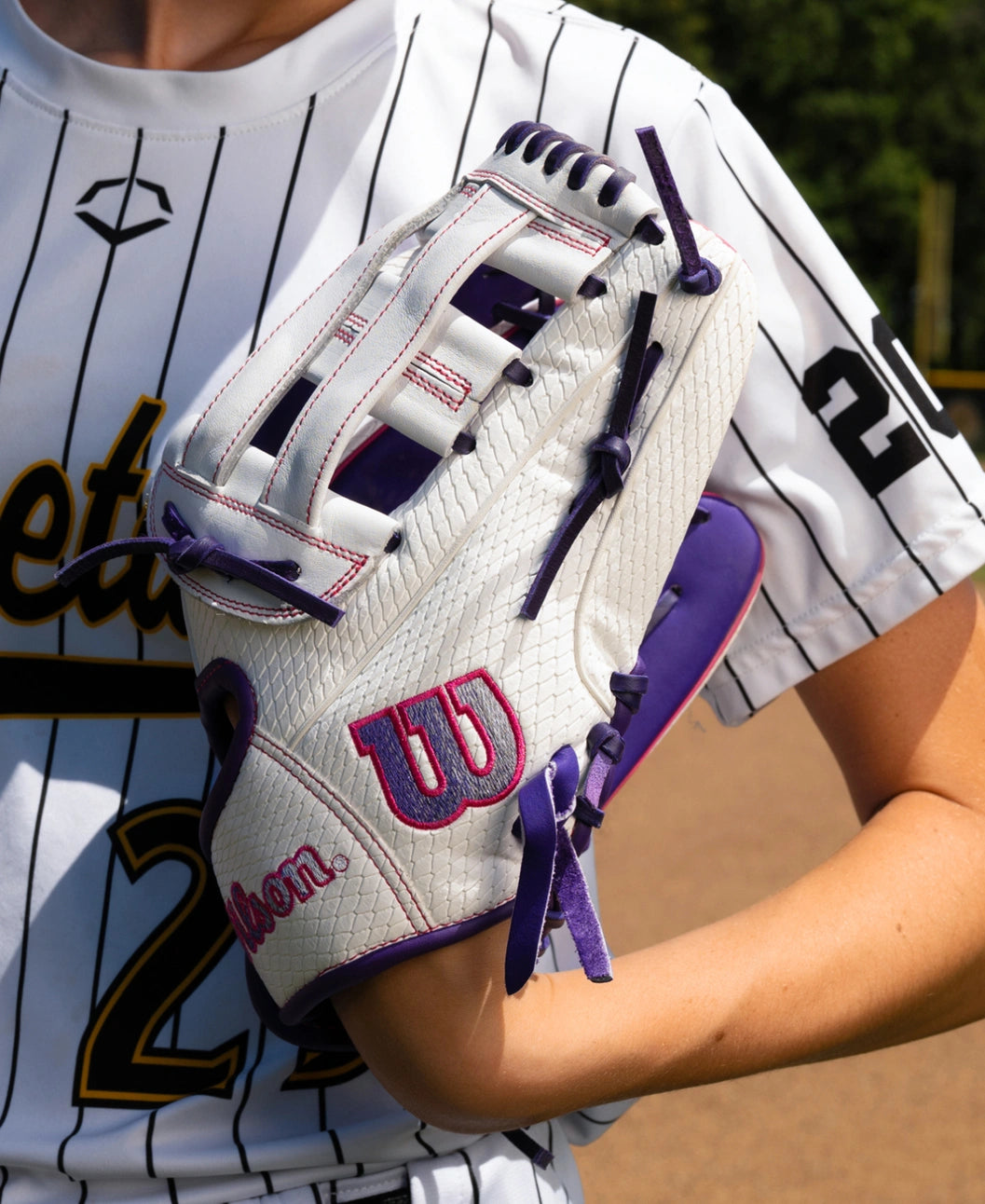 A player wearing a white pinstriped jersey holds a Wilson A1000 FP1712SS 12″ Fastpitch Glove, a full grain leather glove with a Dual Post Web, purple lacing, and purple trim.