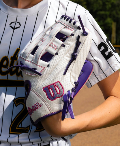 A player wearing a white pinstriped jersey holds a Wilson A1000 FP1712SS 12″ Fastpitch Glove, a full grain leather glove with a Dual Post Web, purple lacing, and purple trim.