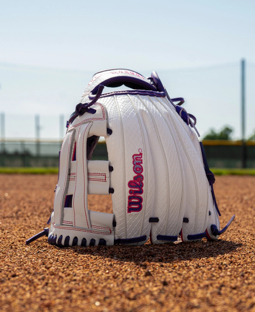 A white and purple Wilson A1000 FP1712SS 12″ Fastpitch Glove with a dual post web sitting on a dirt field.