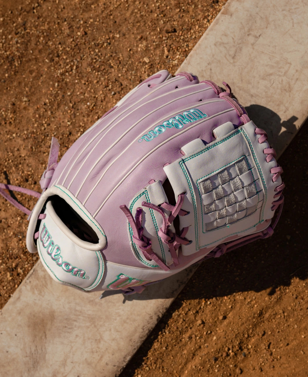 A light purple and white Wilson A1000 P12 12″ Fastpitch Glove: WBG03401RTP12 with teal stitching and lacing resting on a dugout bench.