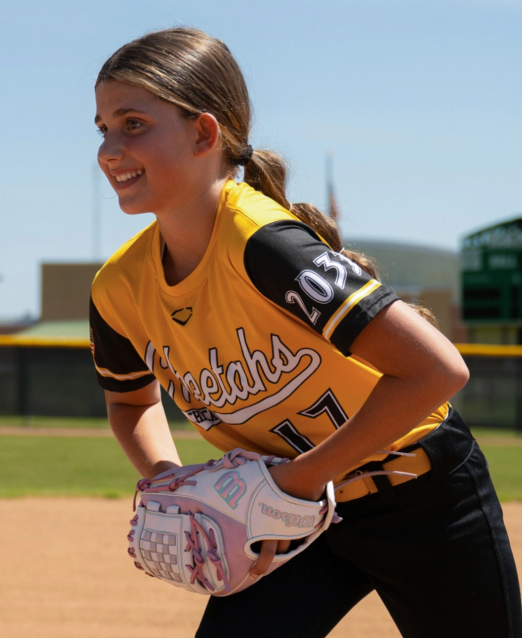 A softball player in a yellow and black jersey stands on a field holding a white and pink Wilson A1000 P12 12″ Fastpitch Glove: WBG03401RTP12.