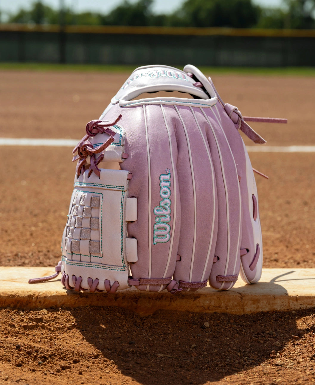 Light purple Wilson A1000 P12 12″ Fastpitch Glove with a checkerboard web, sitting on a baseball field base.
