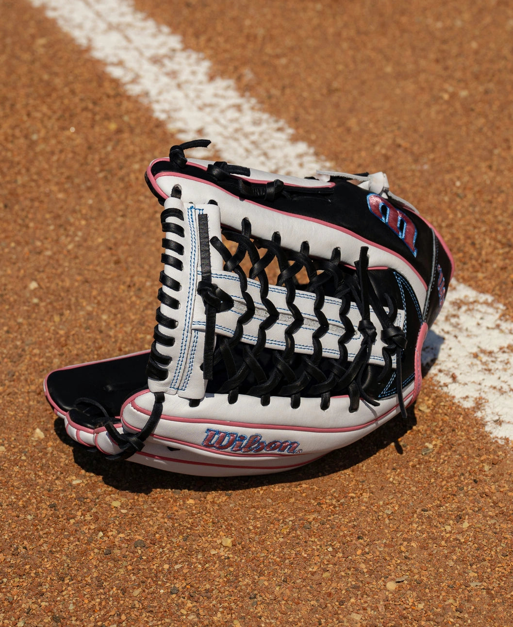 Wilson A1000 T125 12.5″ Fastpitch Glove in black, white, and pink resting on a dirt field with a white foul line.
