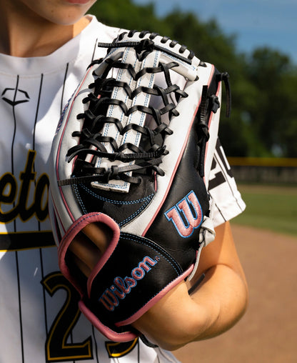 A black and white Wilson A1000 T125 12.5″ Fastpitch Glove: WBG03501RTT125 with pink piping and blue stitching, worn on a player’s hand.