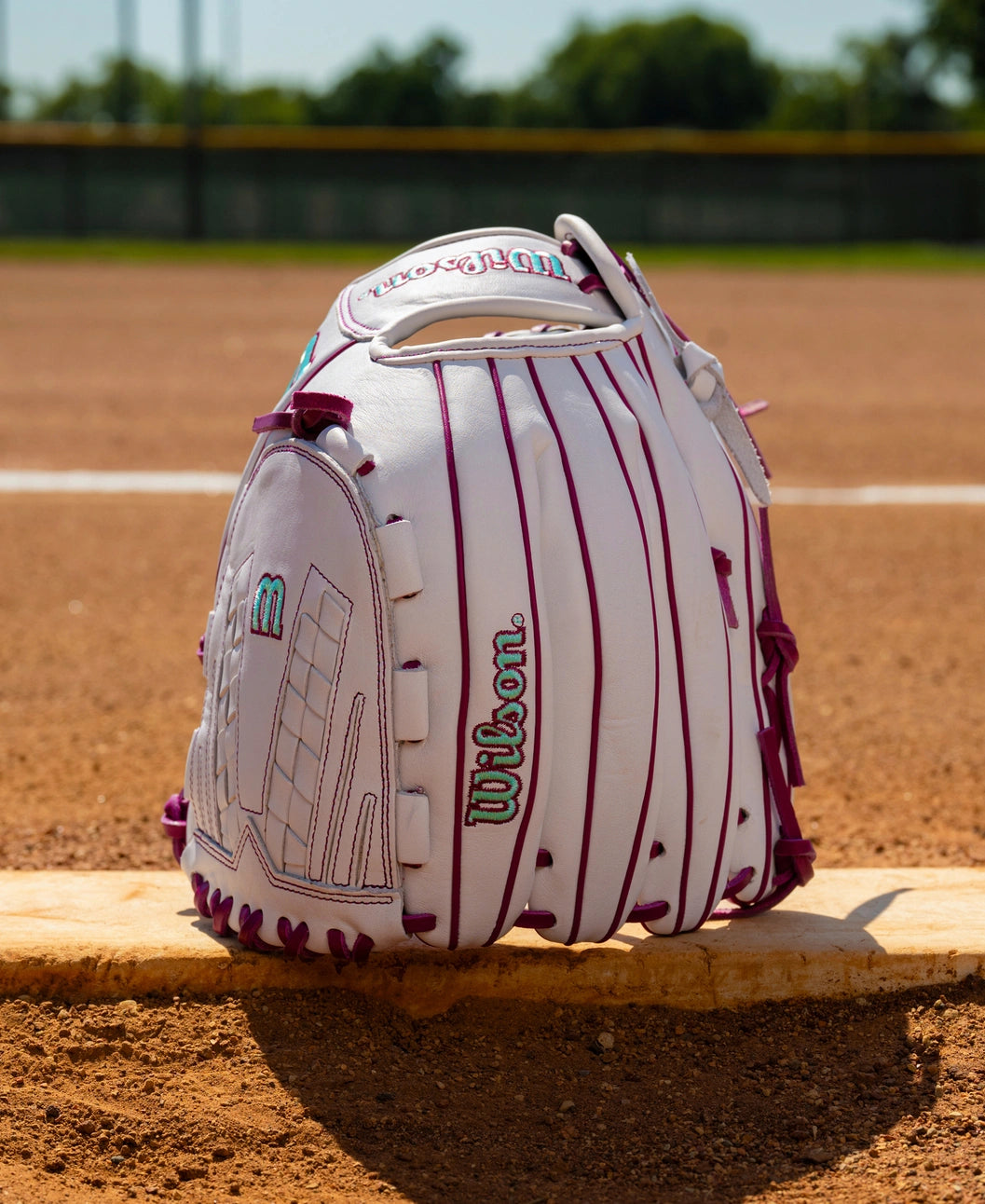 White Wilson A1000 V125 12.5″ Fastpitch Glove with purple lacing and teal detailing, sitting on a pitcher’s plate on a dirt field.