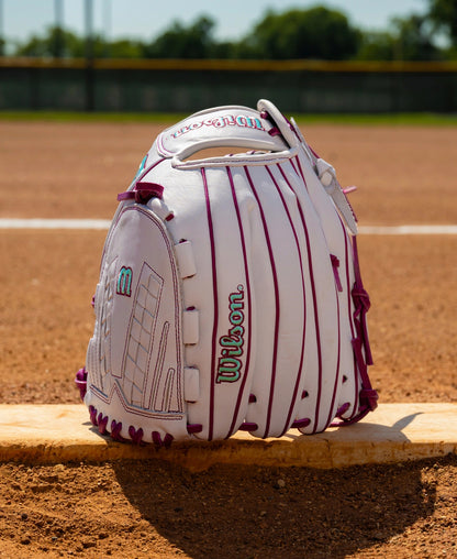 White Wilson A1000 V125 12.5″ Fastpitch Glove with purple lacing and teal detailing, sitting on a pitcher’s plate on a dirt field.