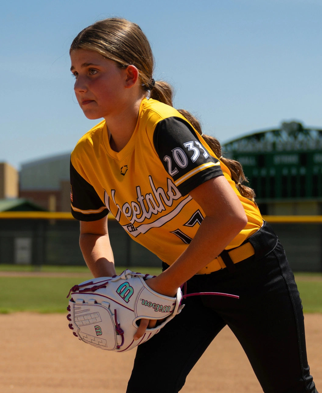 A softball player in a yellow and black jersey stands in a ready position on a field, holding a Wilson A1000 V125 12.5″ Fastpitch Glove: WBG03601RTV125.