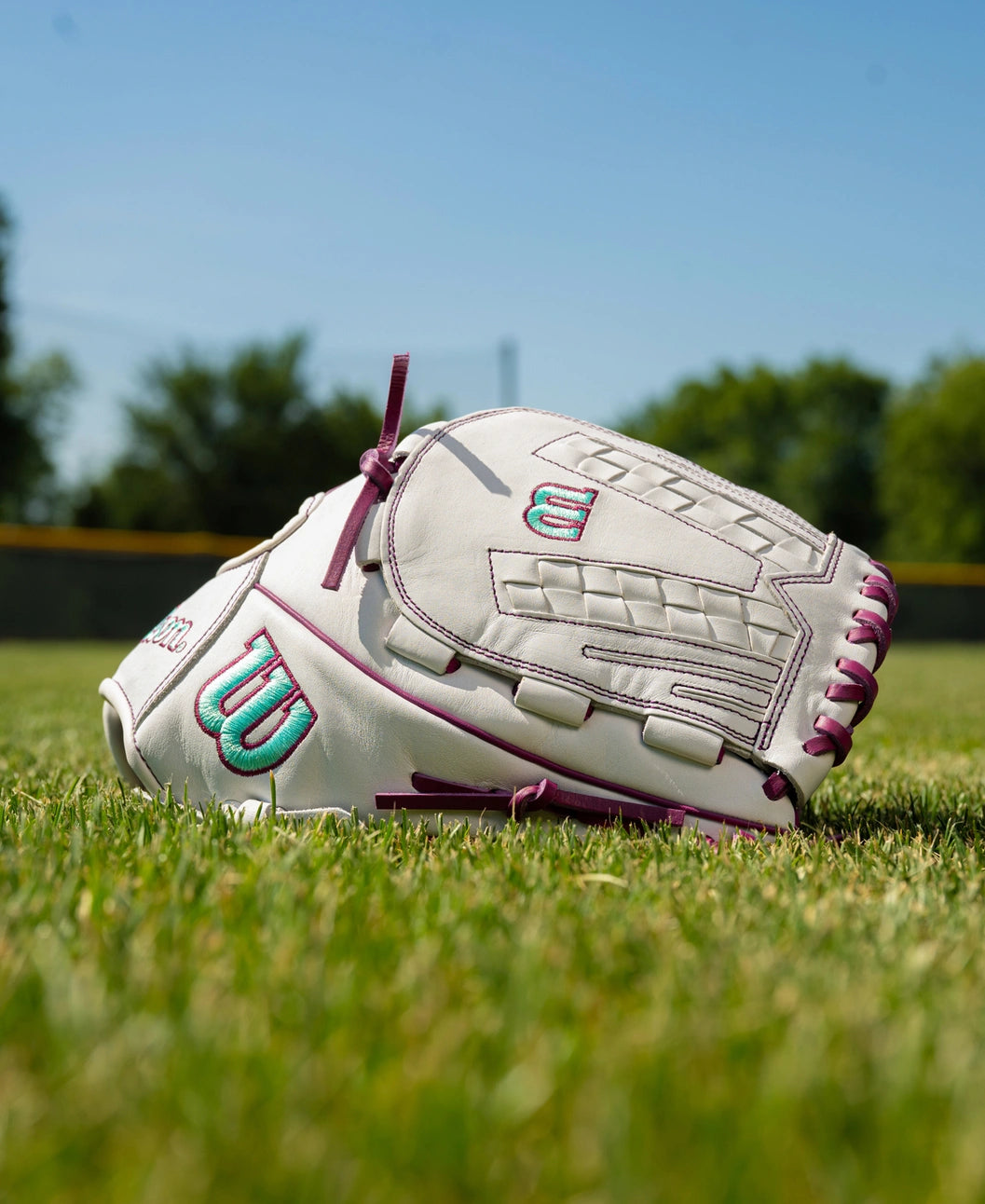 White 12.5″ Wilson A1000 V125 12.5″ Fastpitch Glove: WBG03601RTV125 with purple lacing and teal and purple logos, resting on a grass field.