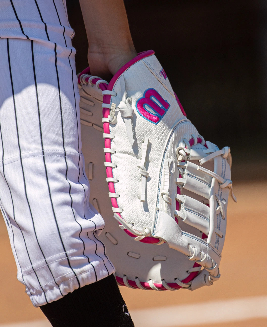 A white Wilson A1000 FP1620SS 12.5″ Fastpitch First Base Mitt with pink trim and a pink logo, held by a player in pinstriped softball pants.