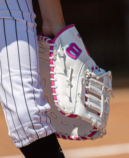 A white Wilson A1000 FP1620SS 12.5″ Fastpitch First Base Mitt with pink trim and a pink logo, held by a player in pinstriped softball pants.