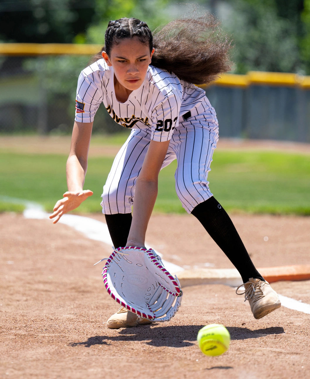 A softball player in a pinstriped uniform reaches down to field a ground ball with a Wilson A1000 FP1620SS 12.5″ Fastpitch First Base Mitt.
