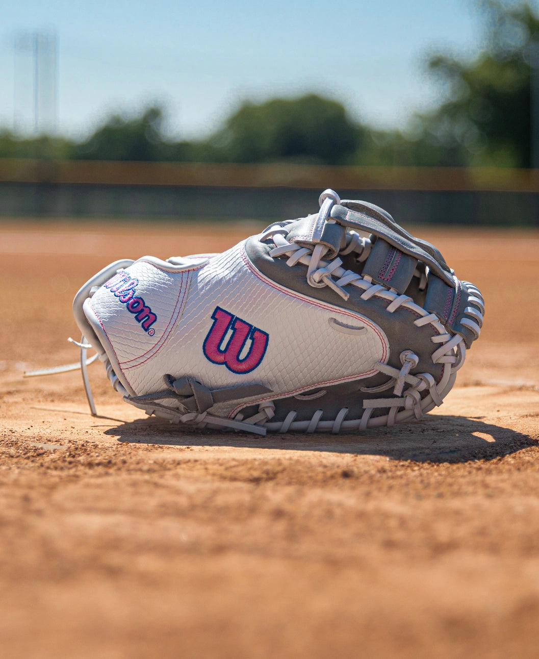 White and grey Wilson A1000 FPCM33 33″ Fastpitch Catcher’s Mitt with pink accents sitting on a dirt softball field.