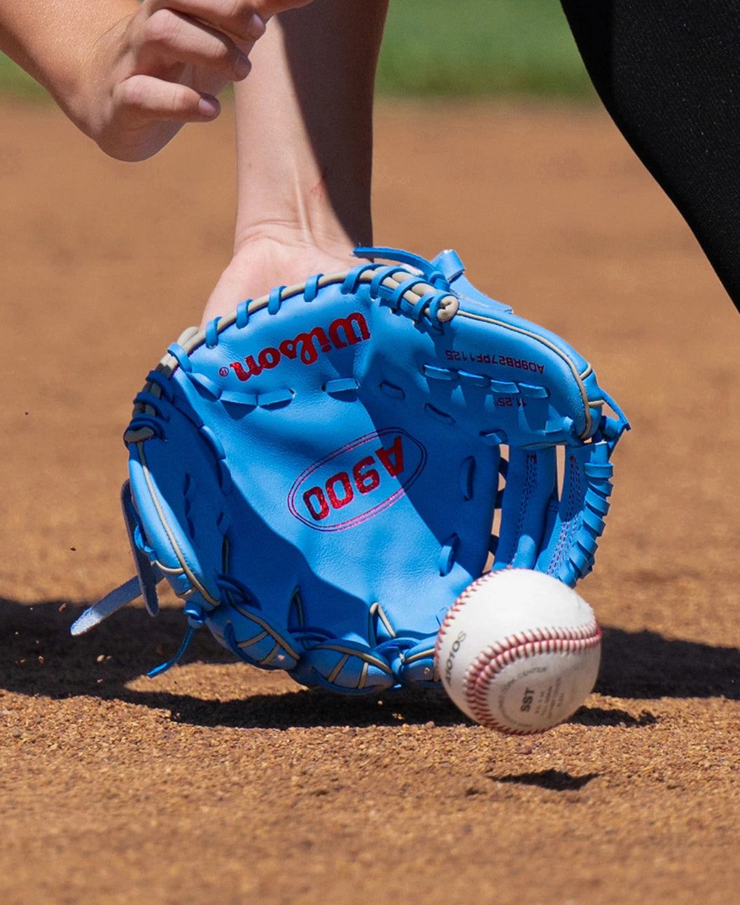 A blue Wilson A900 11.25″ Baseball Glove: WBG03901RT1125 scoops a baseball off a dirt field.