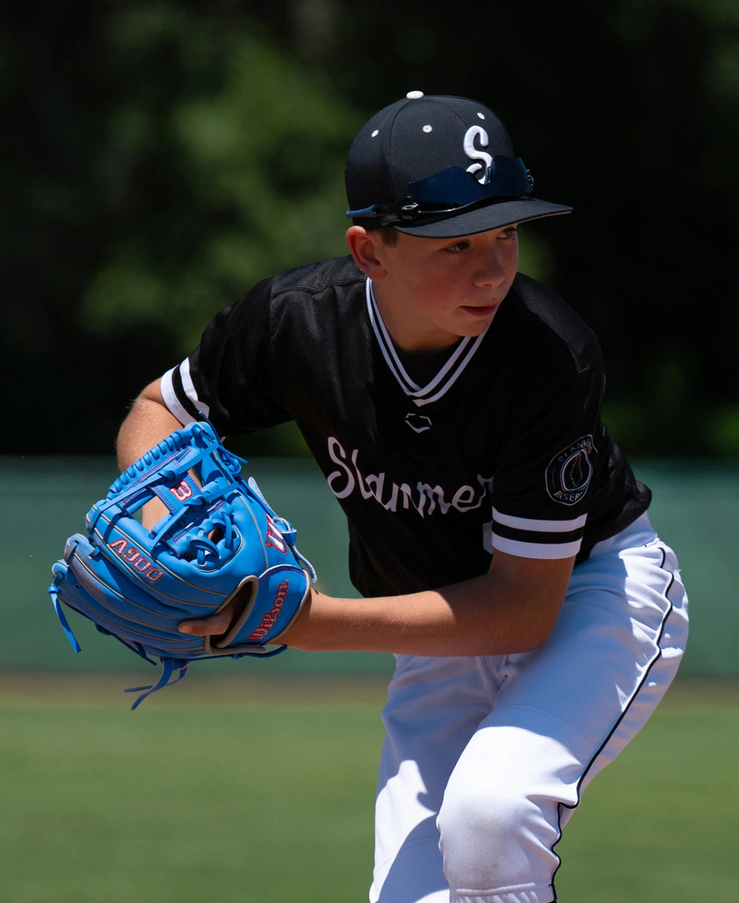 A young baseball player in a black uniform and cap holds a bright blue Wilson A900 11.25″ Baseball Glove: WBG03901RT1125 while preparing to throw.