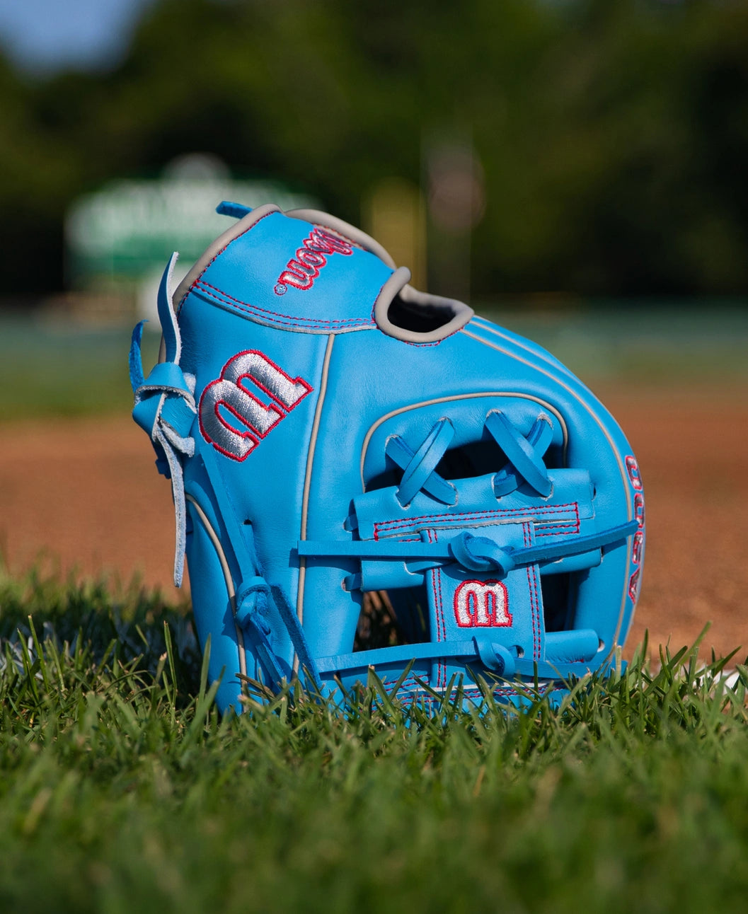 Light blue Wilson A900 11.25″ Baseball Glove: WBG03901RT1125 with red stitching and logos sitting in the grass on a baseball field.