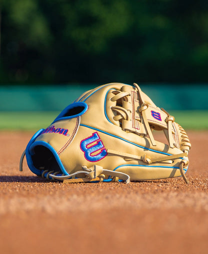 Wilson A900 11.5″ Baseball Glove: WBG04002RT115 in tan with bright blue trim and pink embroidered logos resting on a dirt infield.