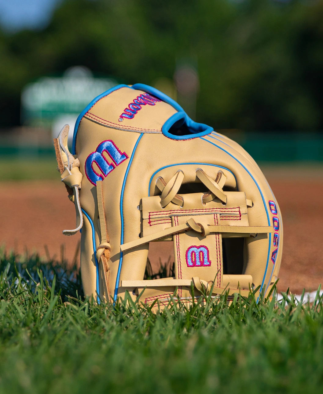 A tan Wilson A900 11.5″ Baseball Glove: WBG04002RT115 with blue piping and pink embroidered logos resting on a baseball field.