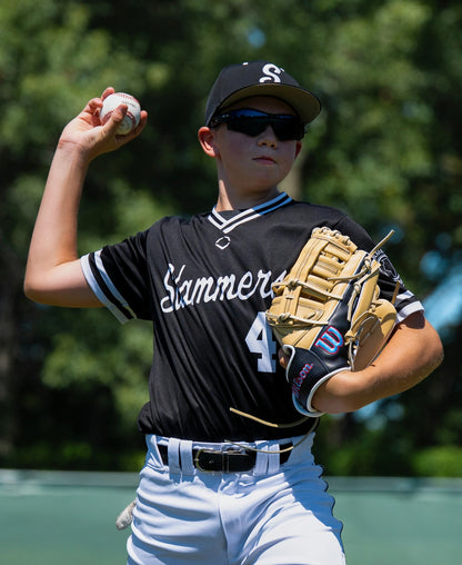 A young baseball player wearing a black uniform and sunglasses prepares to throw a ball, wearing a Wilson A900 12″ Baseball Glove: WBG04101RT12 crafted from full grain leather on their left hand.