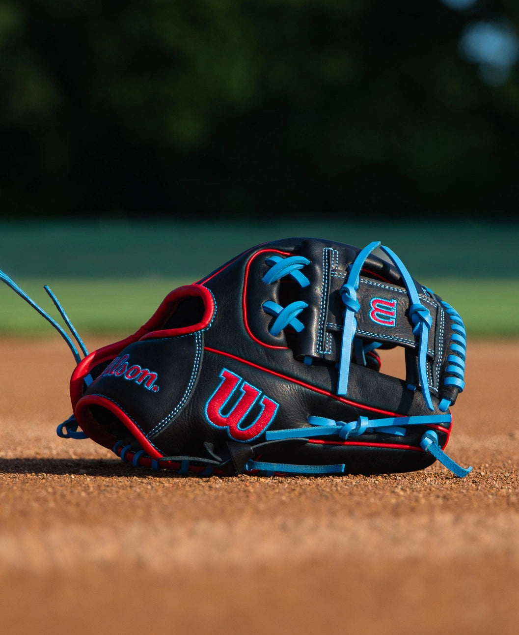 A black Wilson A700 11.5″ Baseball Glove: WBG04401RT115 with bright blue lacing and red trim resting on a dirt field.