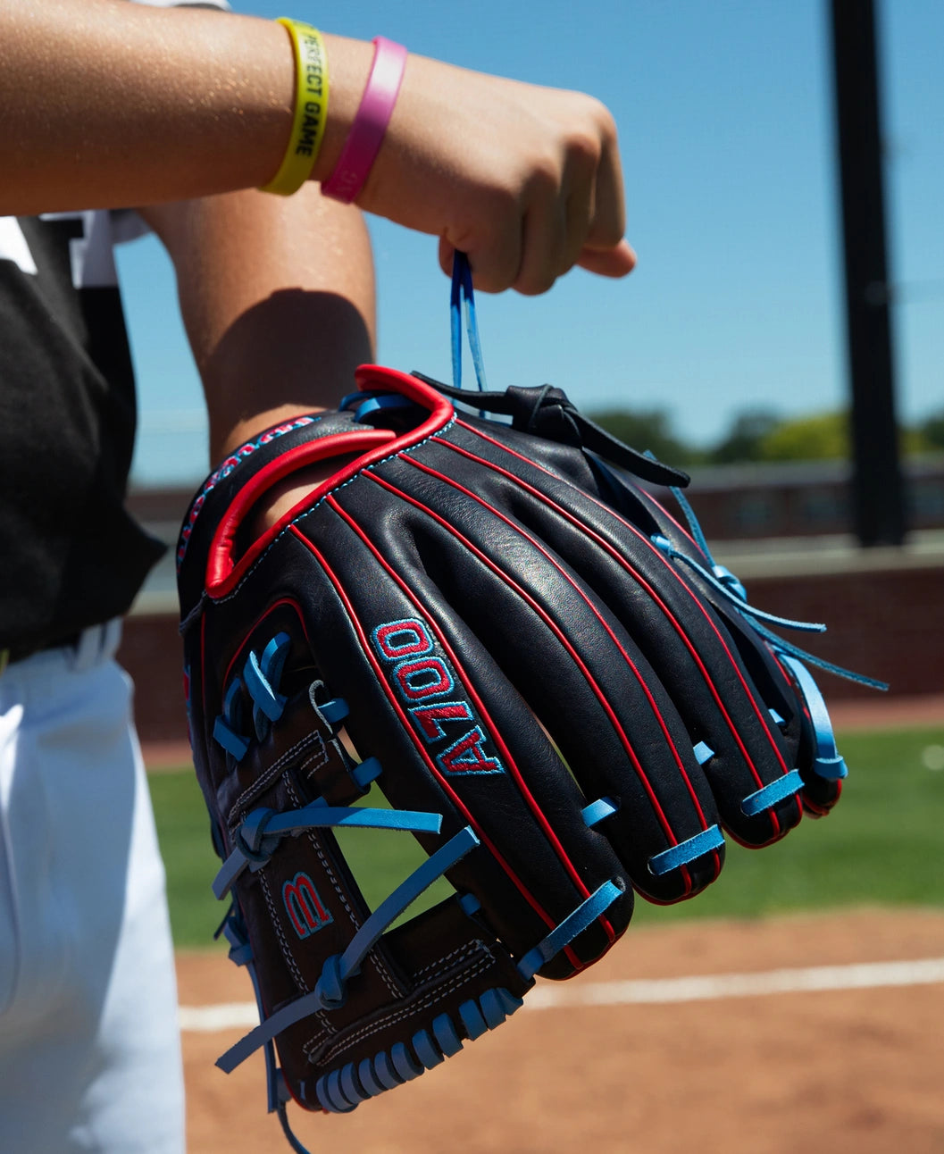 A person holds a black Wilson A700 11.5″ Baseball Glove: WBG04401RT115 with blue laces and red piping on a baseball field.