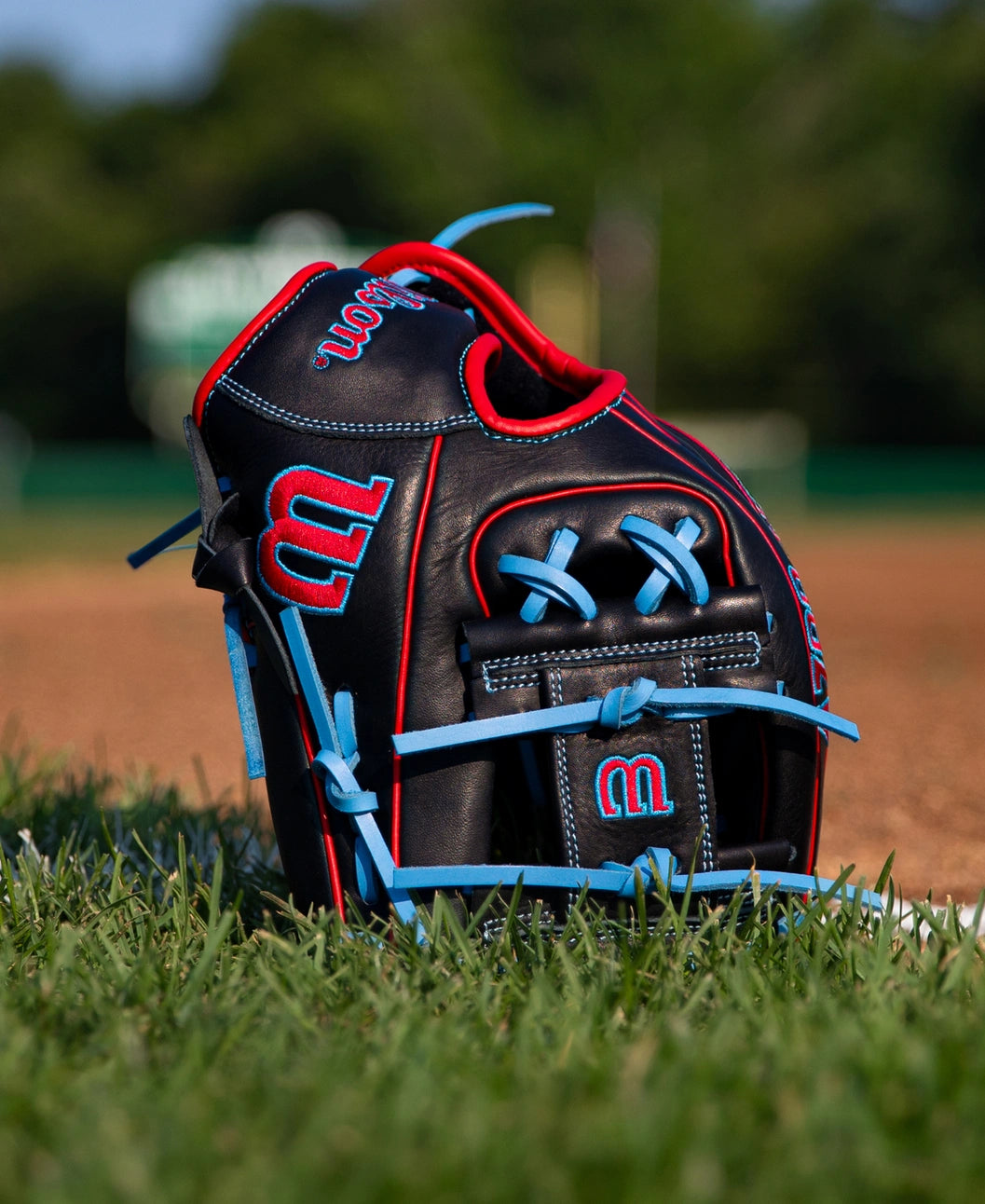 Wilson A700 11.5″ Baseball Glove: WBG04401RT115 made of black full grain leather with bright red and light blue trim, sitting on the grass of a baseball field.