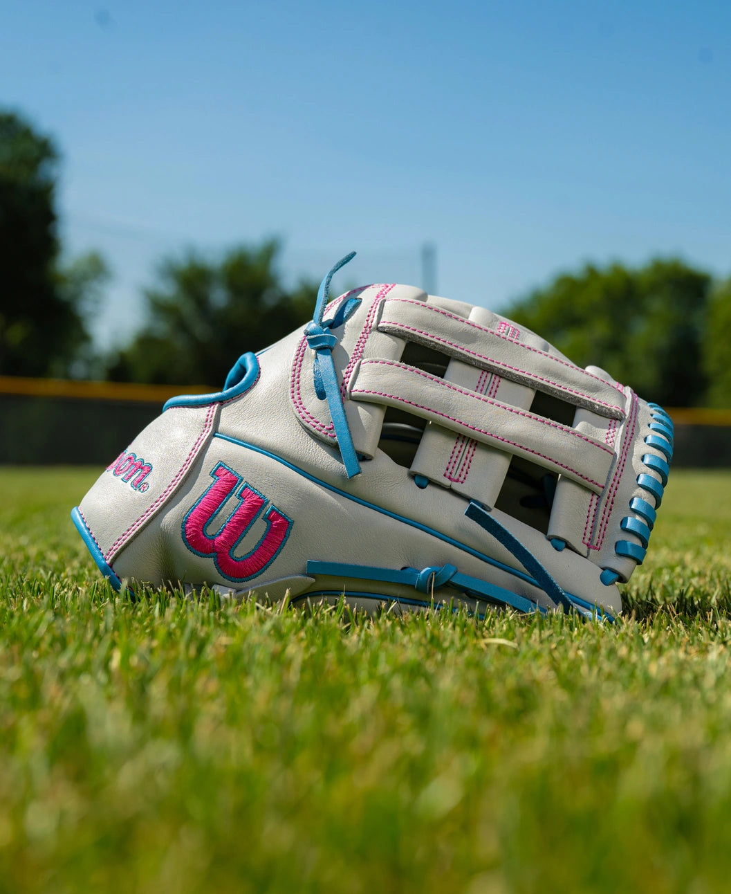 A light gray Wilson A700 12″ Fastpitch Glove: WBG04901RT12 with bright pink and blue accents, crafted from full grain leather, resting on a grass field.