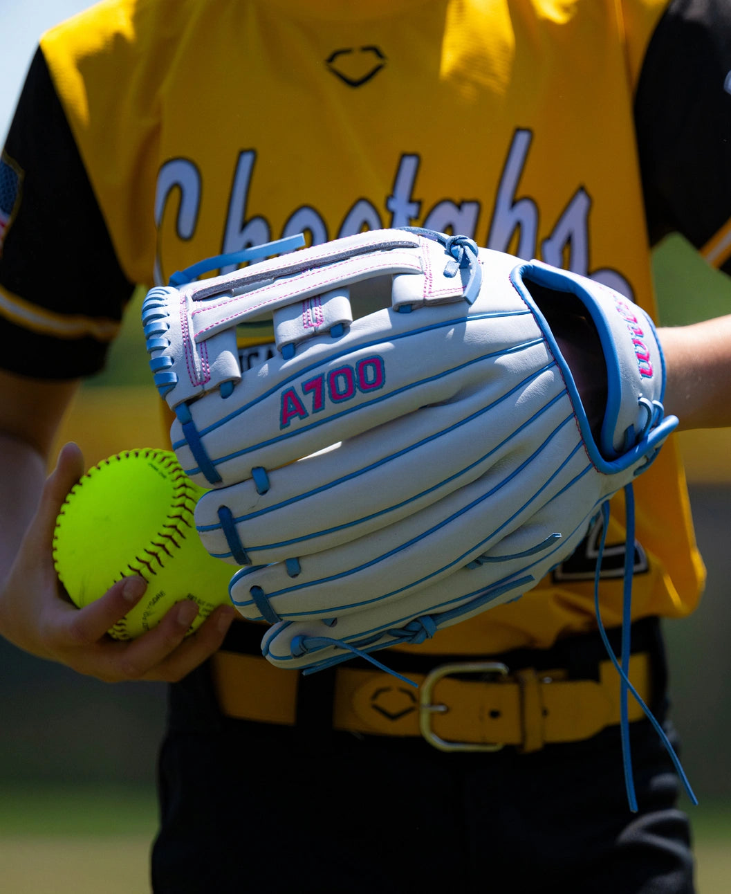 A person in a yellow uniform holds a light blue Wilson A700 12″ Fastpitch Glove: WBG04901RT12 and a yellow softball.