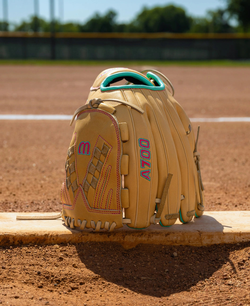 Wilson A700 12.5″ Fastpitch Glove: WBG05001RT125 in tan full grain leather with teal and pink accents, resting on a pitcher’s plate on a dirt infield.