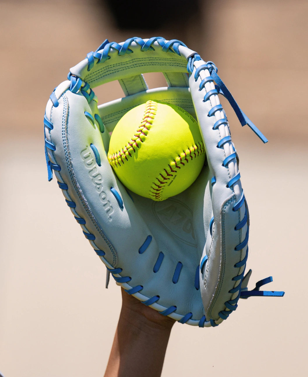 A light-colored Wilson A700 33″ Fastptich Catcher’s Mitt: WBG05101RT33 with bright blue lacing holding a yellow softball in the pocket.