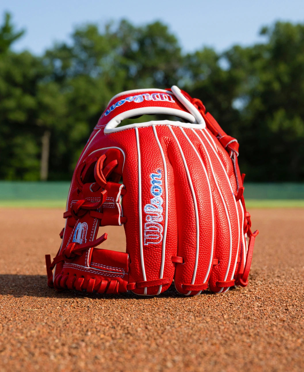 Wilson A1000 PF11SS 11″ Baseball Glove in red with white piping sitting on a dirt field.