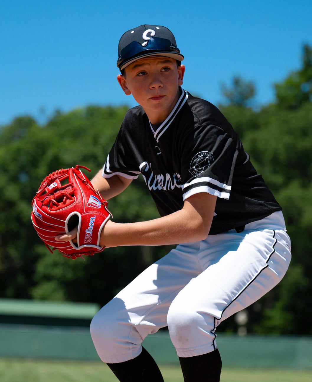 A young baseball player wearing a black and white uniform holds a bright red Wilson A1000 PF11SS 11″ Baseball Glove: WBG05201RT11 in a ready position.