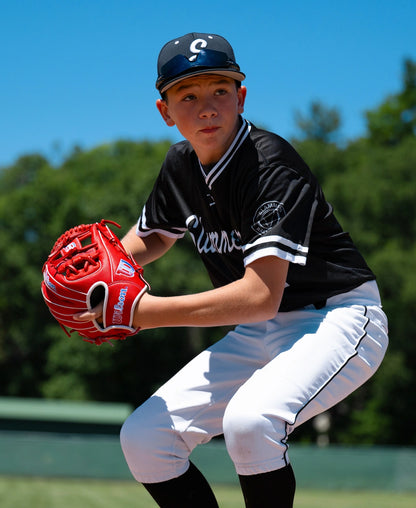 A young baseball player wearing a black and white uniform holds a bright red Wilson A1000 PF11SS 11″ Baseball Glove: WBG05201RT11 in a ready position.
