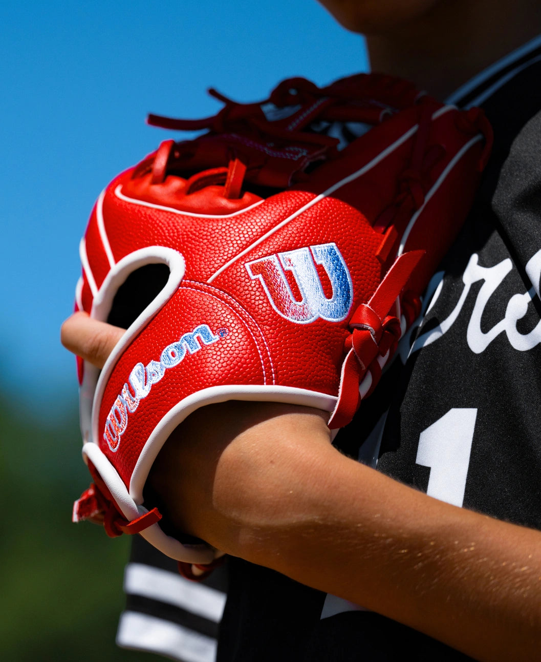 A red Wilson A1000 PF11SS 11″ Baseball Glove: WBG05201RT11 with white trim and blue logo detailing held by a player in a black jersey.