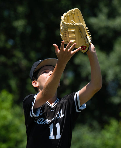 A youth baseball player wearing a uniform reaches up to catch a ball with a Wilson A1000 1892 12.25″ Baseball Glove: WBG05301RT1225.