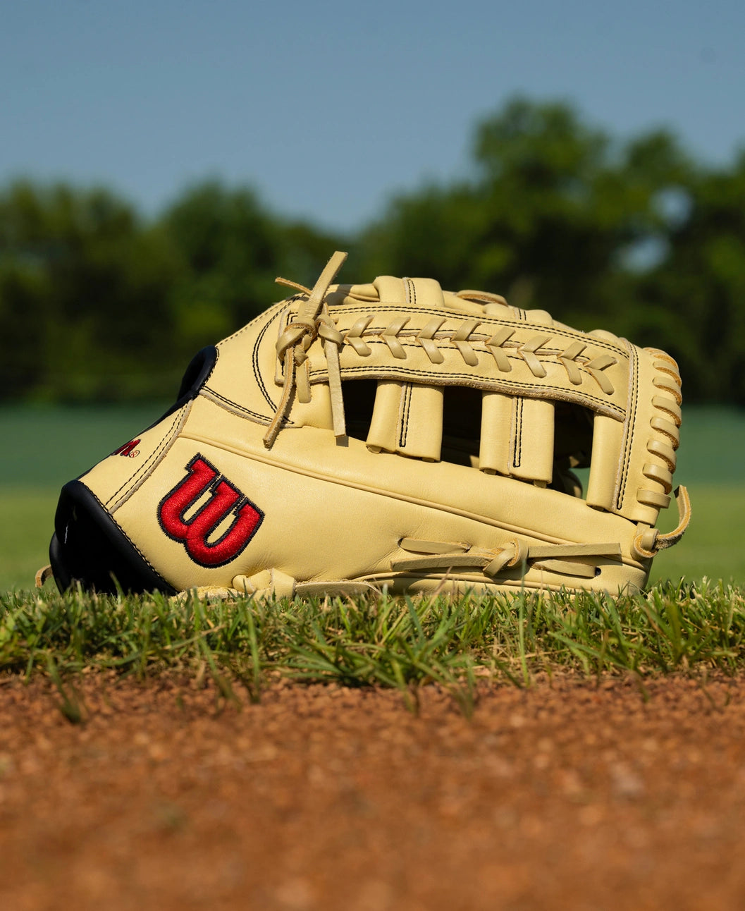 A tan Wilson A1000 1892 12.25″ Baseball Glove: WBG05301RT1225 with black trim and a red logo resting on a baseball field.