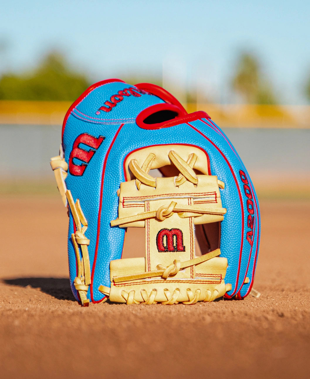 A Wilson A2000 PF88SS 11.25″ Baseball Glove (WBG05402RT1125) by Wilson, in blue and tan with red stitching and Pedroia Fit Technology, sits upright on a dirt baseball field, with a blurred fence and sky in the background.