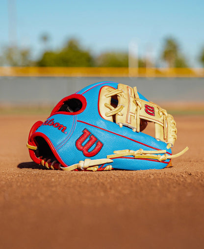A Wilson A2000 PF88SS 11.25″ Baseball Glove (WBG05402RT1125) by Wilson, in blue and tan with red accents and Pedroia Fit Technology, rests on the dirt of a baseball field under a clear sky.