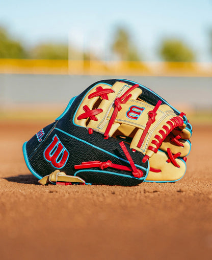 A Wilson A2000 DP15SS 11.5″ Baseball Glove: WBG05501RT115 by Wilson, featuring red laces and blue trim and crafted with the Pedroia Fit for infielders, rests on the dirt of a baseball field as a blurred fence and trees fade into the background.