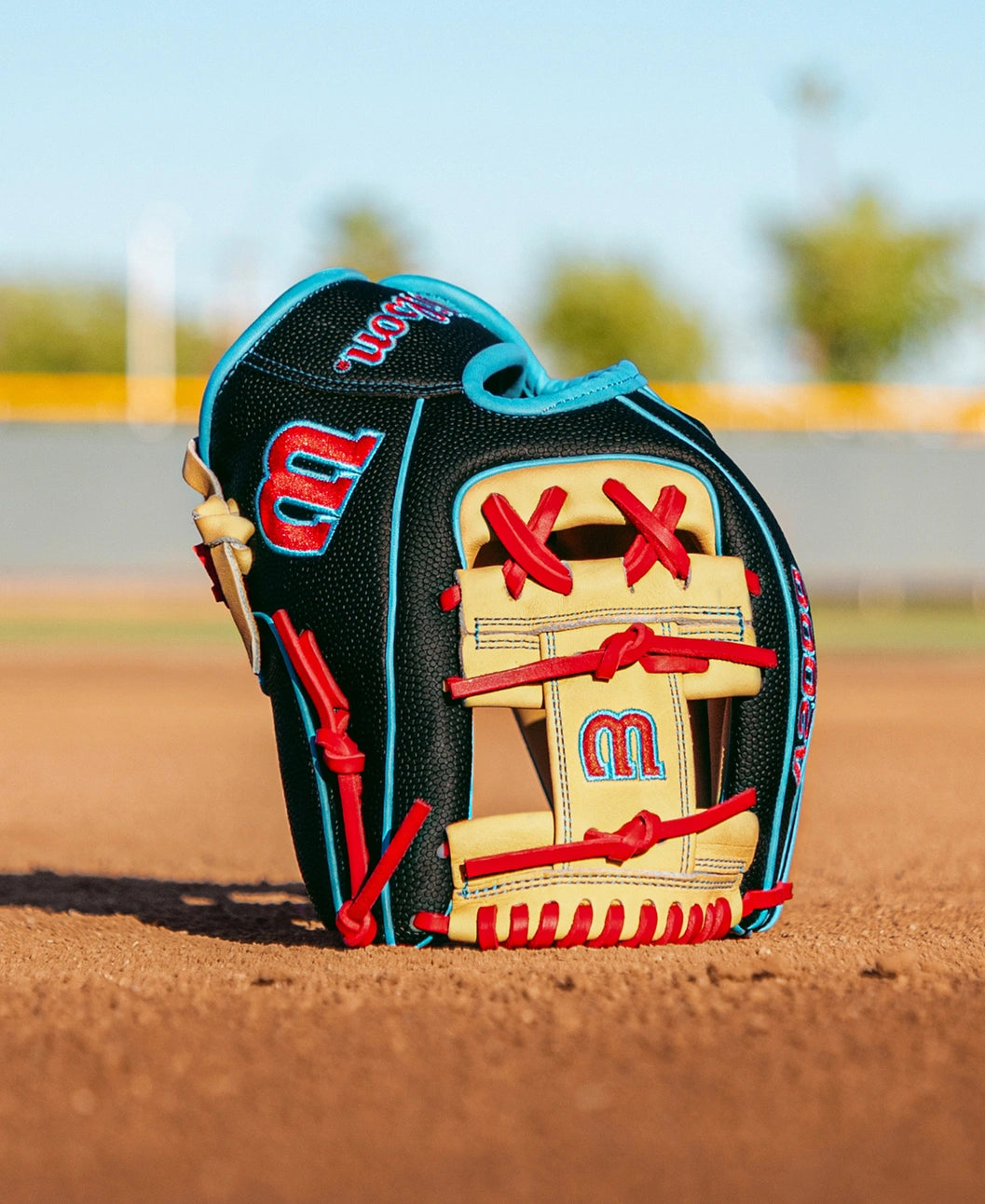 A Wilson A2000 DP15SS 11.5″ Baseball Glove (WBG05501RT115) by Wilson, featuring red, blue, and cream colors, sits upright on a dirt infield with a blurred field and trees in the background.