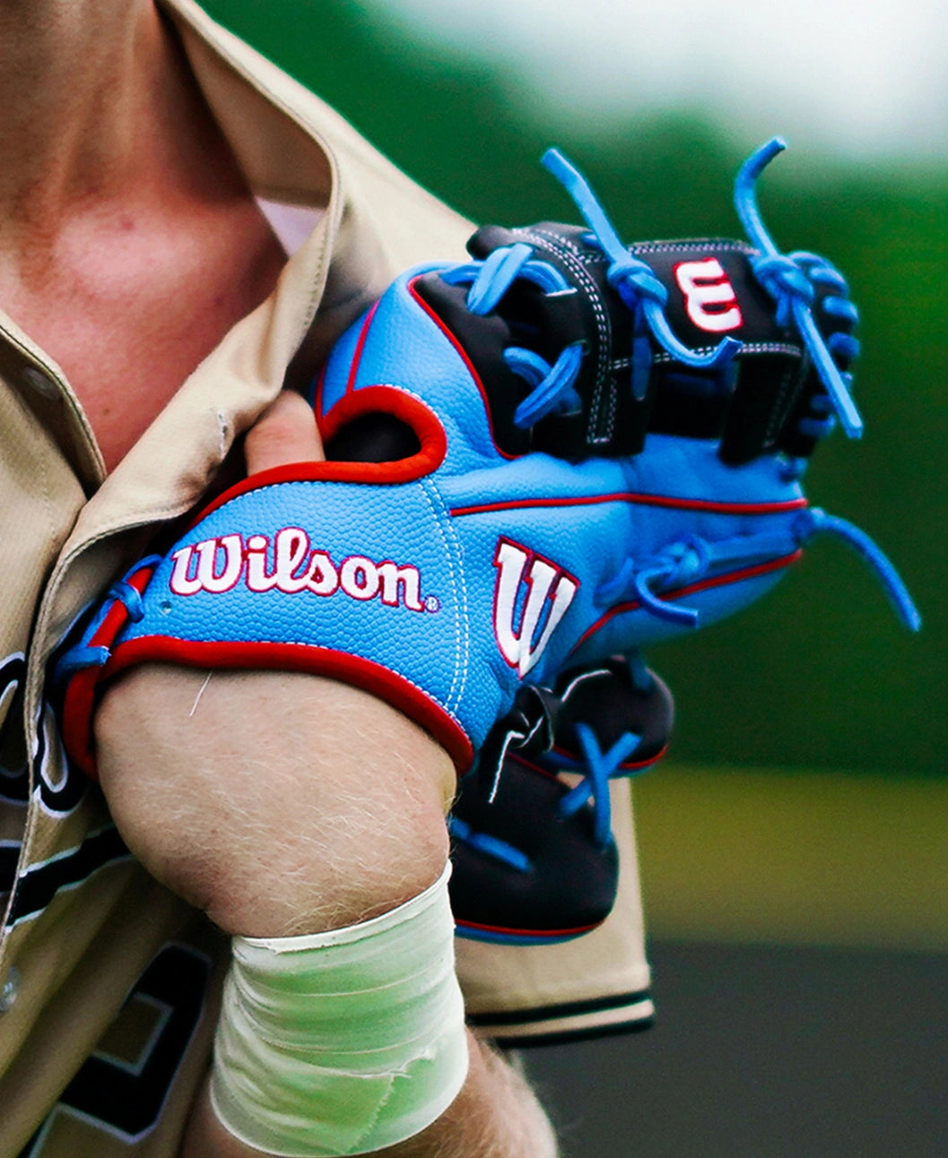 A baseball player in a tan jersey holds a Wilson A2000 1786SS 11.5″ Baseball Glove (WBG05601RT115) with blue and red accents over their shoulder, while white sports tape is wrapped around their wrist.