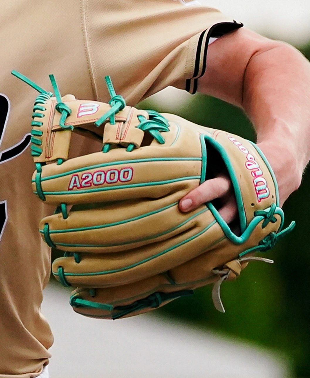 A close-up shot captures a person wearing the Wilson A2000 1975 11.75″ Baseball Glove (WBG05701RT1175) by Wilson—a tan glove accented with teal laces and an extra deep pocket—poised to catch a ball. The 11.75” infield glove stands out as the individual sports a beige jersey with black striped sleeves.