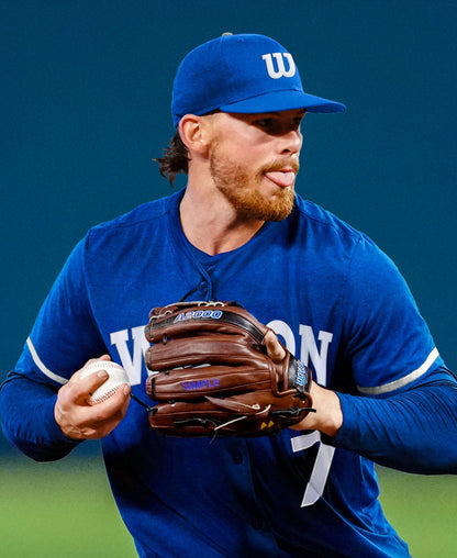 A baseball player in a blue uniform and cap prepares to pitch, gripping a baseball in his right hand and wearing a Wilson A2000 BWJ7GM 11.75″ Bobby Witt Jr. GM Baseball Glove (WBG05801RT1175) on his left hand, tongue out in concentration.