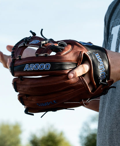 A person stands outdoors beneath a blue sky with trees in the background, wearing a Wilson A2000 BWJ7GM 11.75″ Bobby Witt Jr. GM Baseball Glove (WBG05801RT1175); only the glove and part of the arm are visible.
