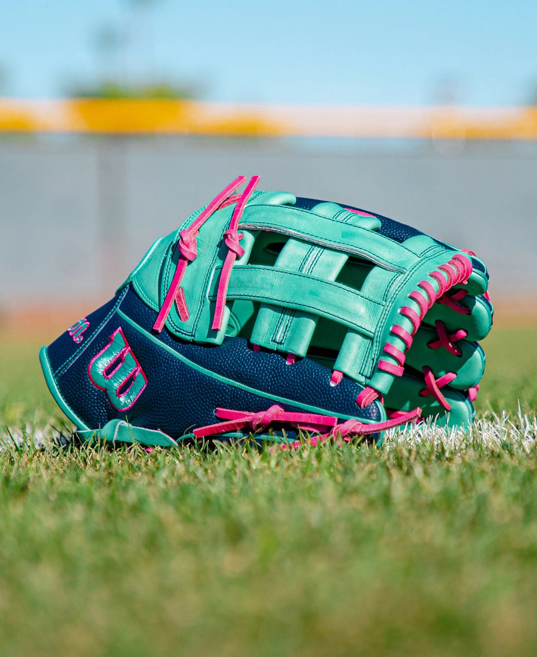 A teal and navy blue Wilson A2000 1750SS 12.5″ Baseball Glove (WBG05901RT125) with pink laces rests on green grass near a white line, while a blurred fence and sky appear in the background. This Wilson glove features a Flex Insert for enhanced comfort and performance.