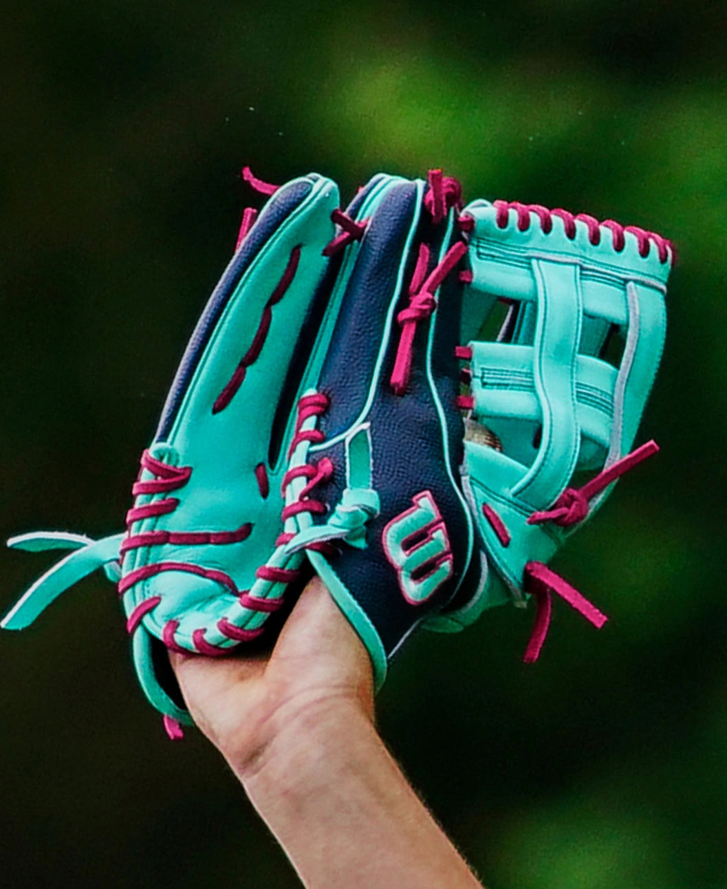 A person holds up a Wilson A2000 1750SS 12.5″ Baseball Glove (WBG05901RT125) in teal and navy blue with pink laces and Flex Insert by Wilson against a blurred green background.